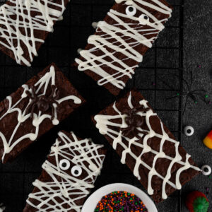 Mehrere rechteckige Halloween-Brownies von oben auf einem schwarzen Kuchengitter: Mumien-Riegel mit weißen Schokoladenbahnen und Zuckeraugen sowie Spinnennetz-Riegel mit dunkler Schoko-Spinne, daneben ein weißes Schälchen mit orange-lila-grünen Zuckerstreuseln und verstreute Zuckeraugen