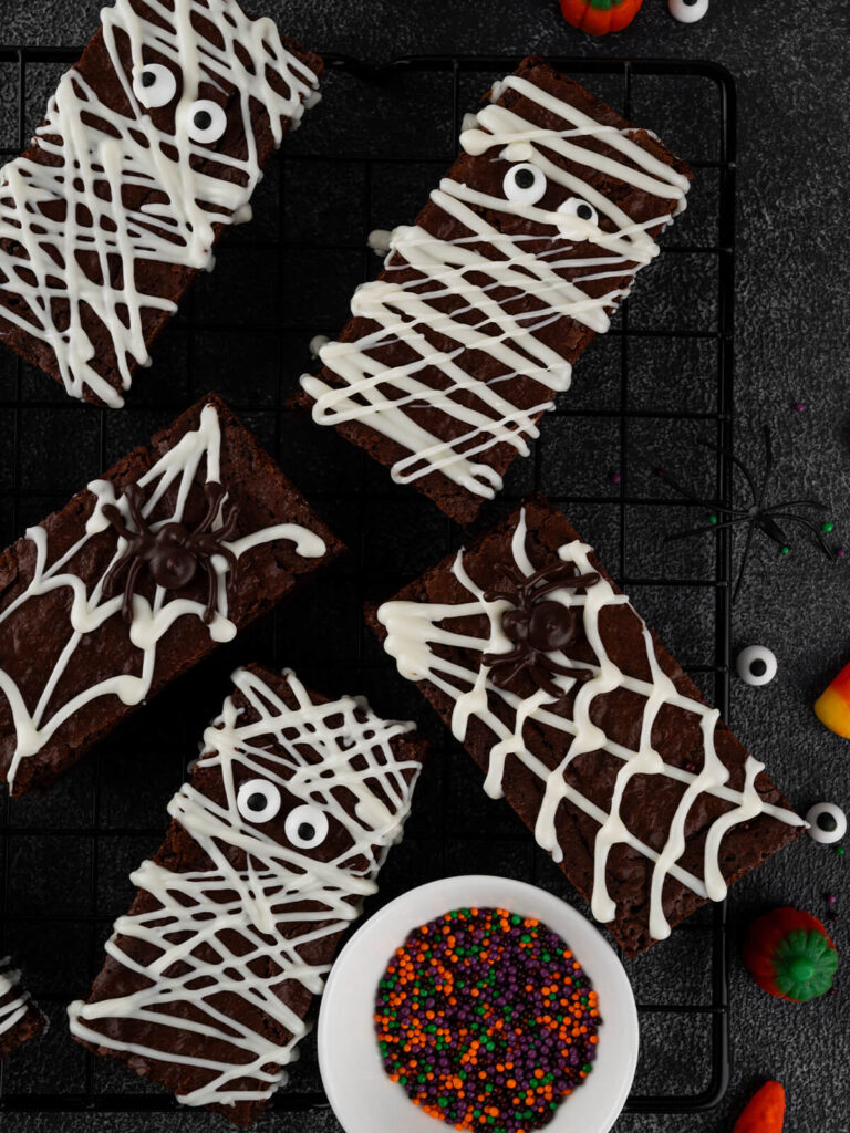 Mehrere rechteckige Halloween-Brownies von oben auf einem schwarzen Kuchengitter: Mumien-Riegel mit weißen Schokoladenbahnen und Zuckeraugen sowie Spinnennetz-Riegel mit dunkler Schoko-Spinne, daneben ein weißes Schälchen mit orange-lila-grünen Zuckerstreuseln und verstreute Zuckeraugen