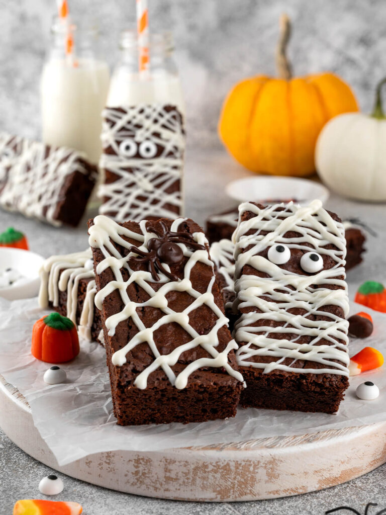 Zwei fertig verzierte Halloween-Brownies auf einem runden Holzbrett mit Backpapier: links ein Spinnennetz-Riegel aus weißer Schokolade mit dunkler Schoko-Spinne, rechts eine Mumie mit weißen Binden und zwei Zuckeraugen, dahinter Milchflaschen mit orange gestreiften Strohhalmen und kleine Kürbisse