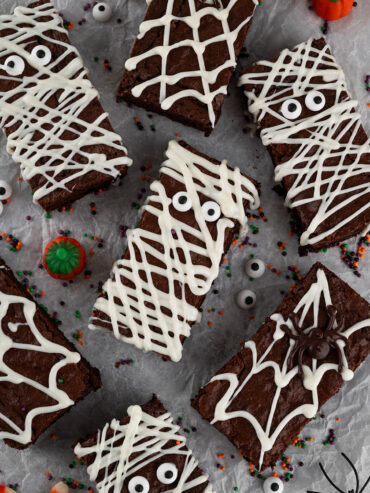 Halloween-Brownies von oben auf hellgrauem Backpapier: in der Mitte eine Mumie mit zwei Zuckeraugen und weißen Schokoladenbahnen, ringsum weitere Mumien-Riegel sowie Spinnennetz-Riegel, darunter einer mit dunkler Schoko-Spinne, dazwischen orange-lila-grüne Zuckerstreusel und verstreute Zuckeraugen