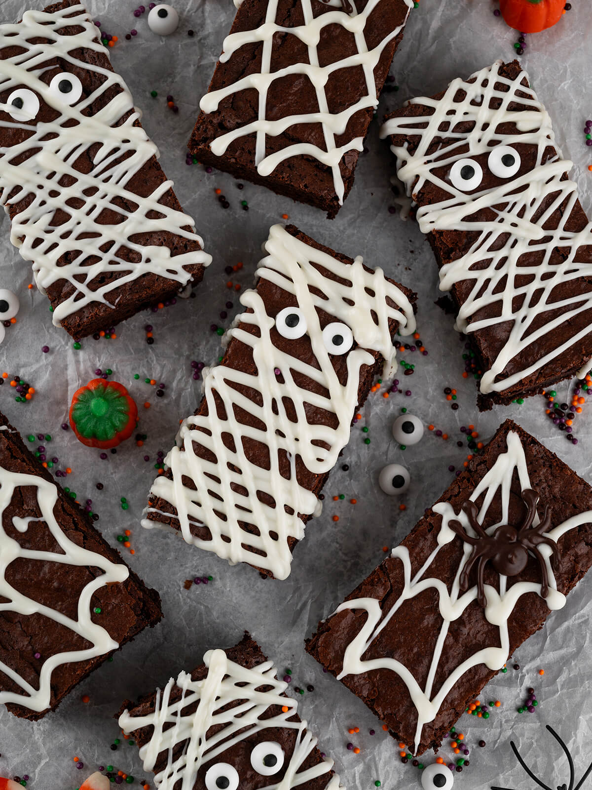 Halloween-Brownies von oben auf hellgrauem Backpapier: in der Mitte eine Mumie mit zwei Zuckeraugen und weißen Schokoladenbahnen, ringsum weitere Mumien-Riegel sowie Spinnennetz-Riegel, darunter einer mit dunkler Schoko-Spinne, dazwischen orange-lila-grüne Zuckerstreusel und verstreute Zuckeraugen
