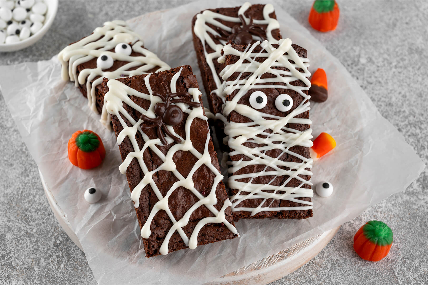 Zwei Halloween-Brownies schräg von der Seite auf weißem Backpapier: vorne ein Spinnennetz-Riegel mit einer dunklen Schoko-Spinne im weißen Netz, daneben eine Mumie mit zwei großen Zuckeraugen und weißen Binden, im Hintergrund ein Schälchen Zuckeraugen und kleine Zuckerkürbisse