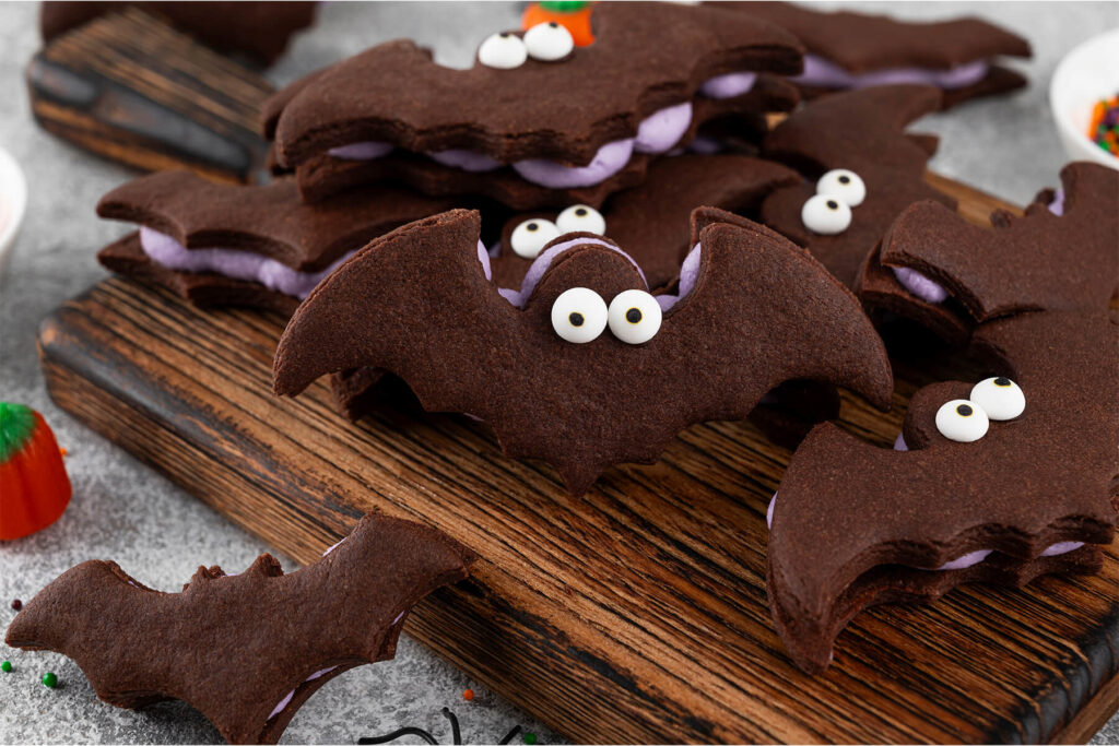 Nahaufnahme dunkler Schoko-Fledermaus-Kekse auf einem dunklen Holzbrett; die Kekse sind paarweise mit hell-lila Buttercreme gefüllt, die seitlich in weichen Wellen hervorquillt, und tragen auf dem Kopf zwei weiße Zuckeraugen mit schwarzen Pupillen; im Vordergrund eine vom Bildrand angeschnittene Fledermaus, links orangefarbene Kürbisbonbons und bunte Streusel auf grauem Steinuntergrund.