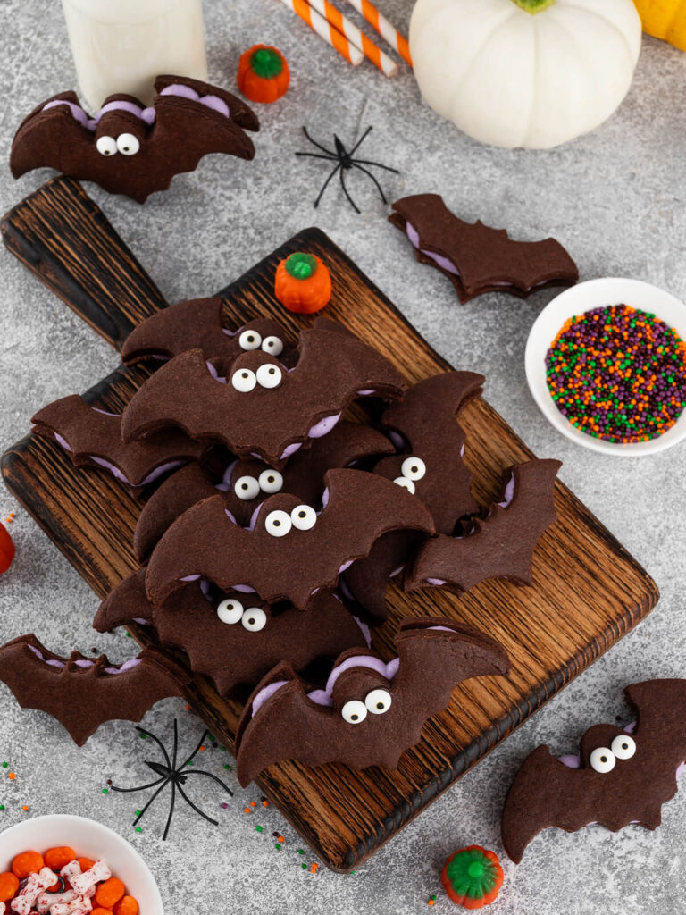 Aufsicht auf ein dunkles Holzbrett voller Schoko-Fledermaus-Sandwichkekse mit hell-lila Buttercreme-Füllung und weißen Zuckeraugen; daneben ein Glas Milch, ein weißer Kürbis, orange gestreifte Papierstrohhalme, ein Schälchen mit orange-lila-grünen Streuseln, Mini-Kürbisbonbons und schwarze Spielzeugspinnen auf grauem Untergrund.