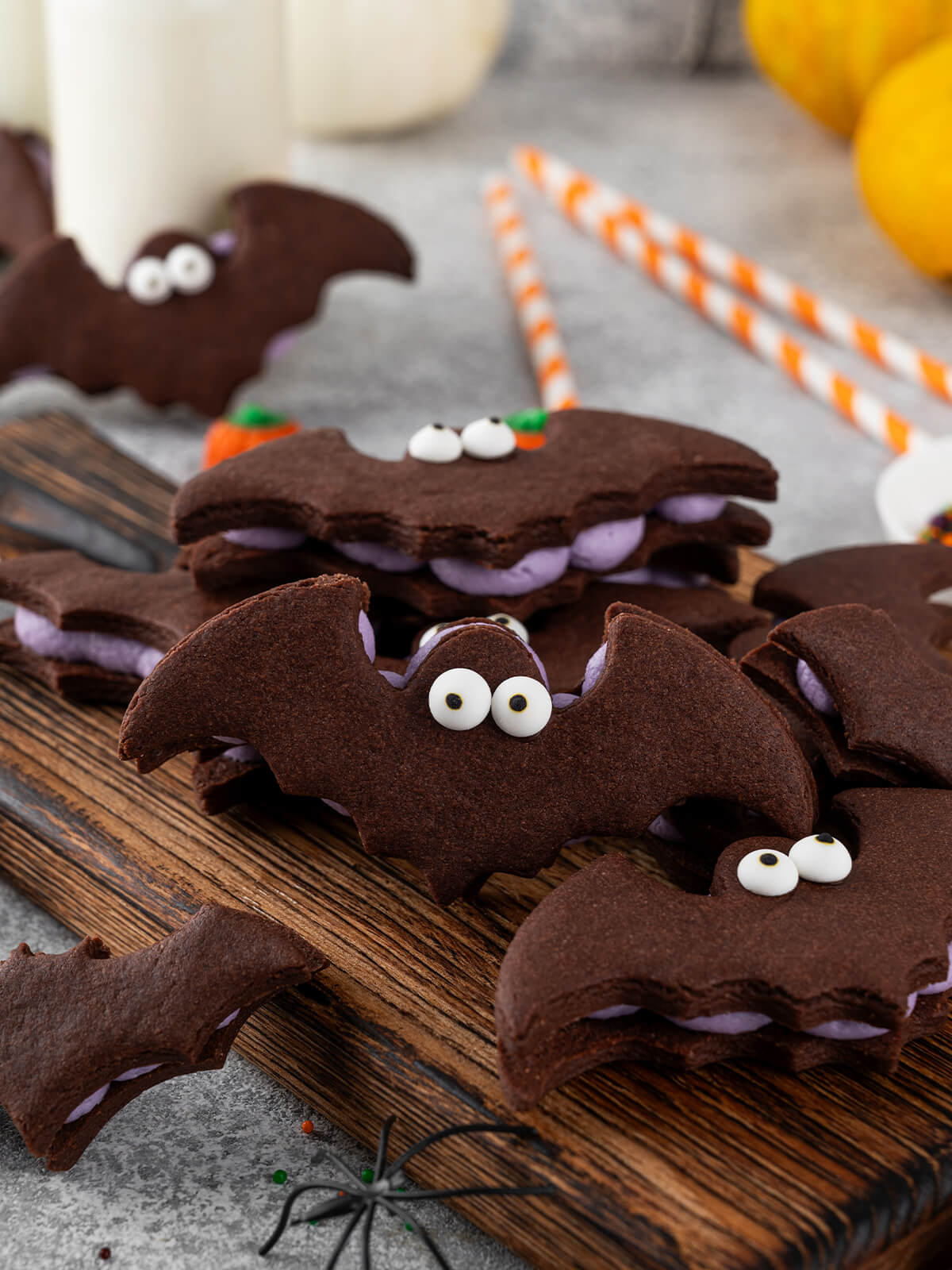 Schräge Nahaufnahme von Schoko-Fledermaus-Sandwichkeksen auf einem Holzbrett; im Fokus eine Fledermaus mit zwei großen weißen Zuckeraugen und hell-lila Buttercreme zwischen den Keksen, im unscharfen Hintergrund ein Milchglas, gelbe Kürbisse und orange gestreifte Papierstrohhalme.