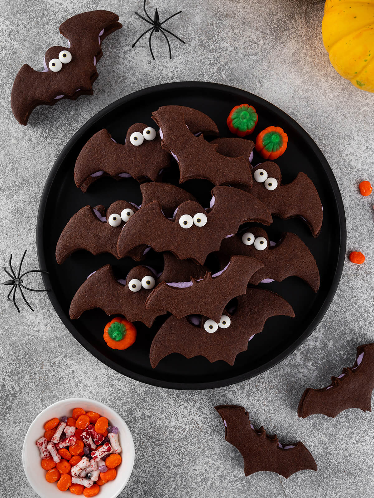 Aufsicht auf einen schwarzen runden Teller mit Schoko-Fledermaus-Sandwichkeksen, jeweils mit hell-lila Buttercreme und zwei weißen Zuckeraugen; ringsum orange und grüne Mini-Kürbisbonbons, schwarze Spielzeugspinnen, oben rechts ein gelber Kürbis und unten links ein weißes Schälchen mit orangefarbenen Bonbons und weißen Knochenstreuseln auf grauem Untergrund