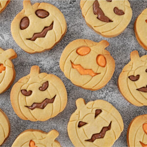 Viele helle Kürbis-Kekse mit geprägten Rillen und Stiel dicht an dicht auf grauem Stein, die ausgestochenen Jack-o-Laterne-Gesichter gefüllt mit brauner Kakao-Buttercreme oder oranger Creme