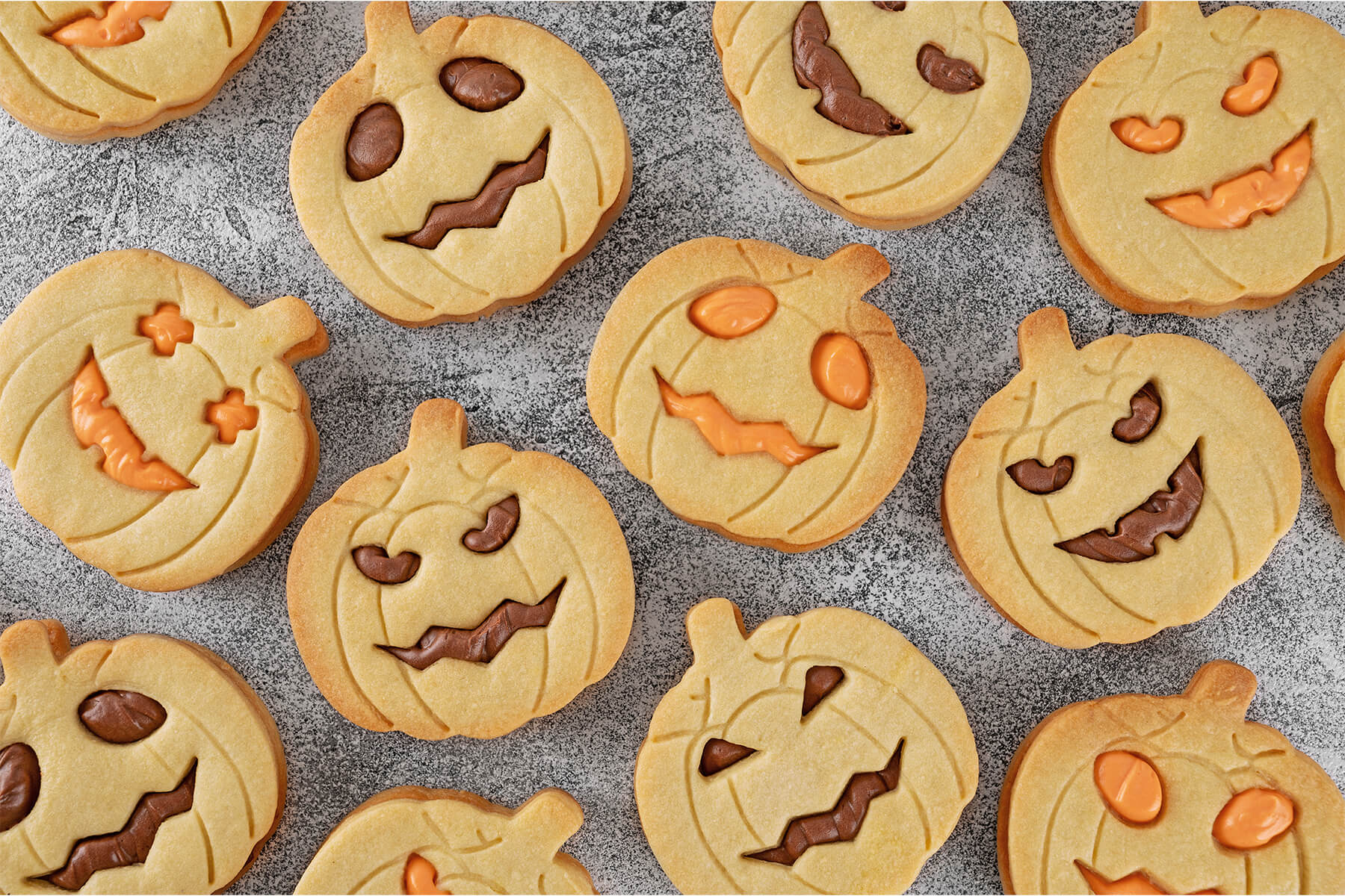 Viele helle Kürbis-Kekse mit geprägten Rillen und Stiel dicht an dicht auf grauem Stein, die ausgestochenen Jack-o-Laterne-Gesichter gefüllt mit brauner Kakao-Buttercreme oder oranger Creme