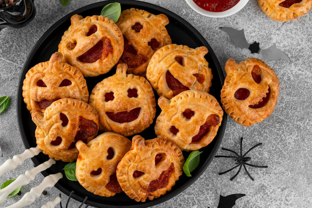 Herzhafte Halloween-Pizza-Pies aus goldbraunem Blätterteig in Kürbisform mit ausgeschnittenen Jack-o-Laterne-Gesichtern auf einem runden schwarzen Teller von oben, die rote Füllung schaut heraus, dazu frisches Basilikum, ein weißes Schälchen mit Tomatensauce, schwarze Fledermäuse, eine Spinne und eine weiße Skeletthand auf hellgrauem Steingrund