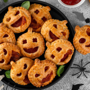Herzhafte Halloween-Pizza-Pies aus goldbraunem Blätterteig in Kürbisform mit ausgeschnittenen Jack-o-Laterne-Gesichtern auf einem runden schwarzen Teller von oben, die rote Füllung schaut heraus, dazu frisches Basilikum, ein weißes Schälchen mit Tomatensauce, schwarze Fledermäuse, eine Spinne und eine weiße Skeletthand auf hellgrauem Steingrund