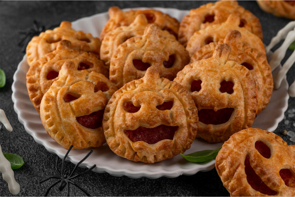 Nahaufnahme goldbrauner Halloween-Pizza-Pies in Kürbisform mit gegabelten Rändern und lachenden Jack-o-Laterne-Gesichtern, die rote Pizzafüllung ist sichtbar, gestapelt auf einem weißen geriffelten Teller vor dunklem Hintergrund mit schwarzer Spinne und Basilikum