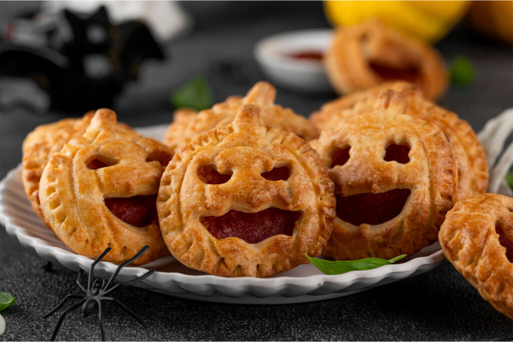 Drei goldbraune Halloween-Pizza-Pies in Kürbisform mit ausgeschnittenen Jack-o-Laterne-Gesichtern und roter Füllung auf einem weißen geriffelten Teller, dunkler Hintergrund mit schwarzer Spinne, unscharfem Saucenschälchen und gelbem Kürbis