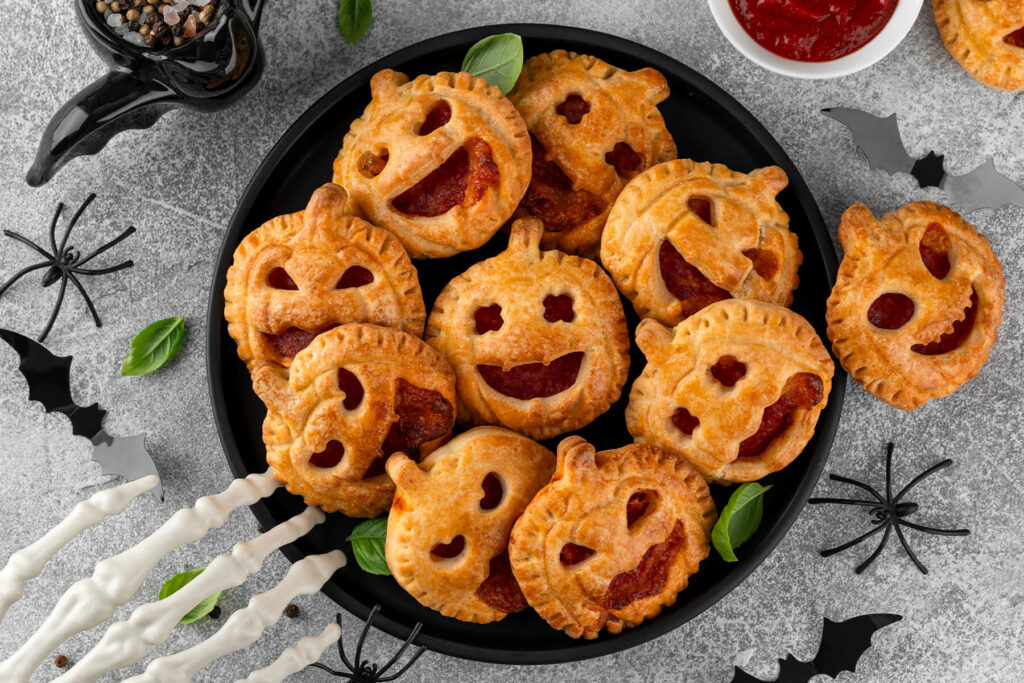 Herzhafte Halloween-Pizza-Pies in Kürbisform von oben auf einem runden schwarzen Teller, jedes Gebäck mit eigenem Jack-o-Laterne-Gesicht und roter Füllung, umringt von frischem Basilikum, schwarzen Fledermäusen, Spinnen und einer weißen Skeletthand auf hellgrauem Steingrund