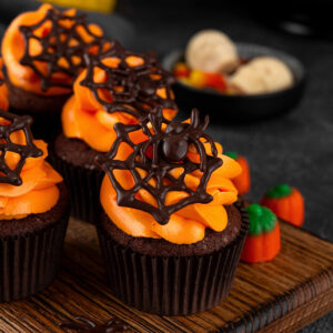 Dunkle Schoko-Cupcakes in braunen Papierförmchen in einer Reihe auf einem Holzbrett, jeder mit breiter oranger Buttercreme-Rosette und einem dunklen Schoko-Spinnennetz, der vordere mit einer Schoko-Spinne, daneben Kürbis-Zuckerdeko