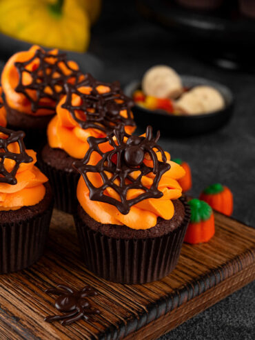 Dunkle Schoko-Cupcakes in braunen Papierförmchen in einer Reihe auf einem Holzbrett, jeder mit breiter oranger Buttercreme-Rosette und einem dunklen Schoko-Spinnennetz, der vordere mit einer Schoko-Spinne, daneben Kürbis-Zuckerdeko