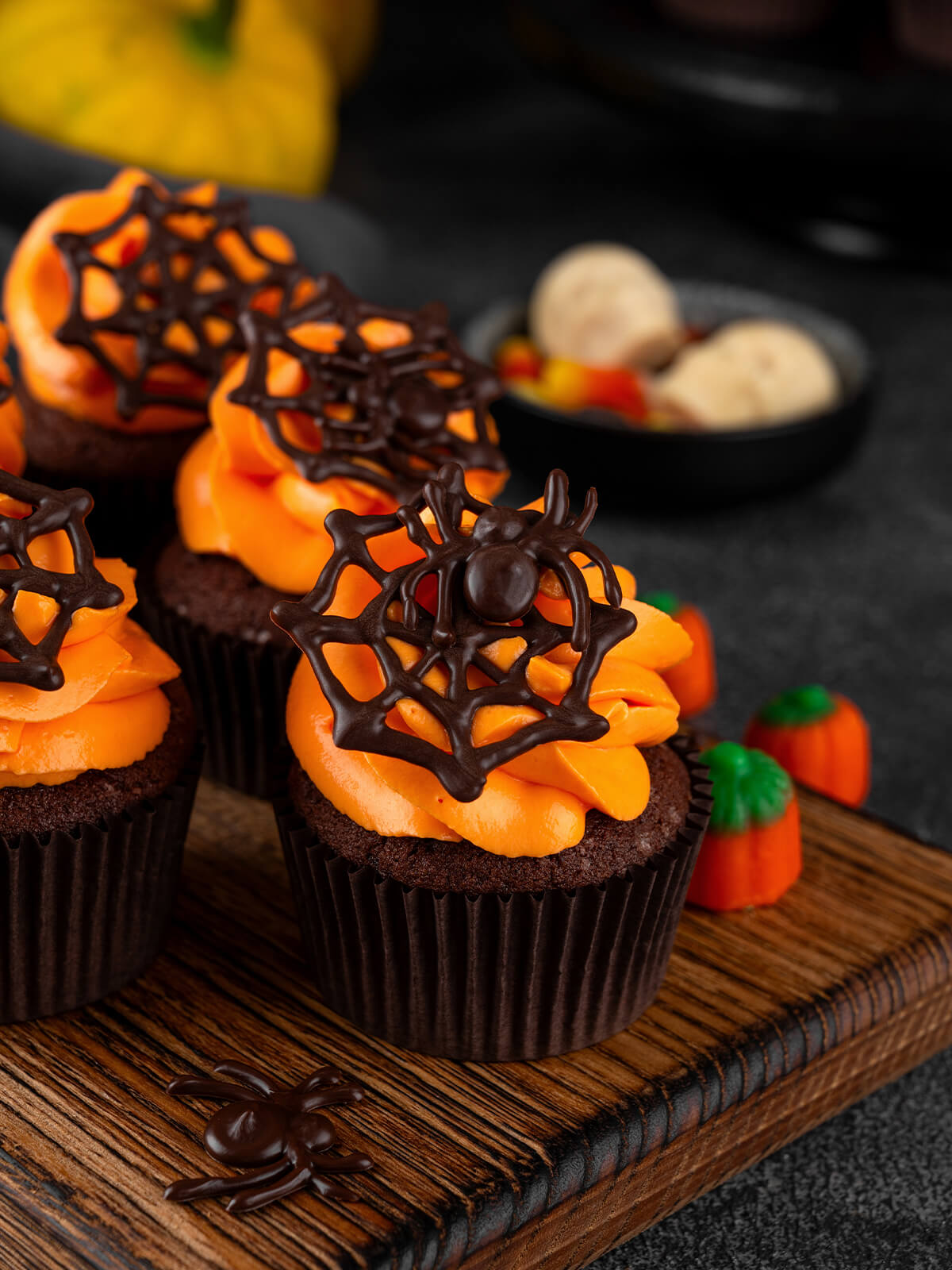 Dunkle Schoko-Cupcakes in braunen Papierförmchen in einer Reihe auf einem Holzbrett, jeder mit breiter oranger Buttercreme-Rosette und einem dunklen Schoko-Spinnennetz, der vordere mit einer Schoko-Spinne, daneben Kürbis-Zuckerdeko