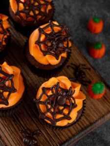 Fertig dekorierte Halloween-Schoko-Cupcakes von oben auf einem Holzbrett, mit oranger Buttercreme und dunklen Schoko-Spinnennetzen, einige mit Schoko-Spinnen, dazu Kürbis-Zuckerdeko und kleine Schoko-Spinnen