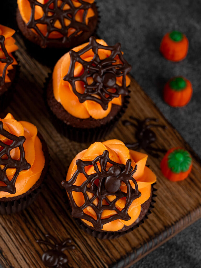 Fertig dekorierte Halloween-Schoko-Cupcakes von oben auf einem Holzbrett, mit oranger Buttercreme und dunklen Schoko-Spinnennetzen, einige mit Schoko-Spinnen, dazu Kürbis-Zuckerdeko und kleine Schoko-Spinnen