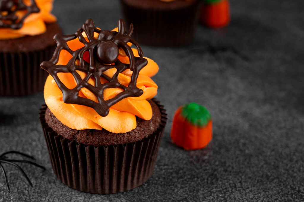 Einzelner dunkler Schoko-Cupcake mit breiter oranger Buttercreme-Rosette und dunklem Schoko-Spinnennetz samt Schoko-Spinne, daneben eine Kürbis-Zuckerfigur, im Hintergrund weitere Cupcakes