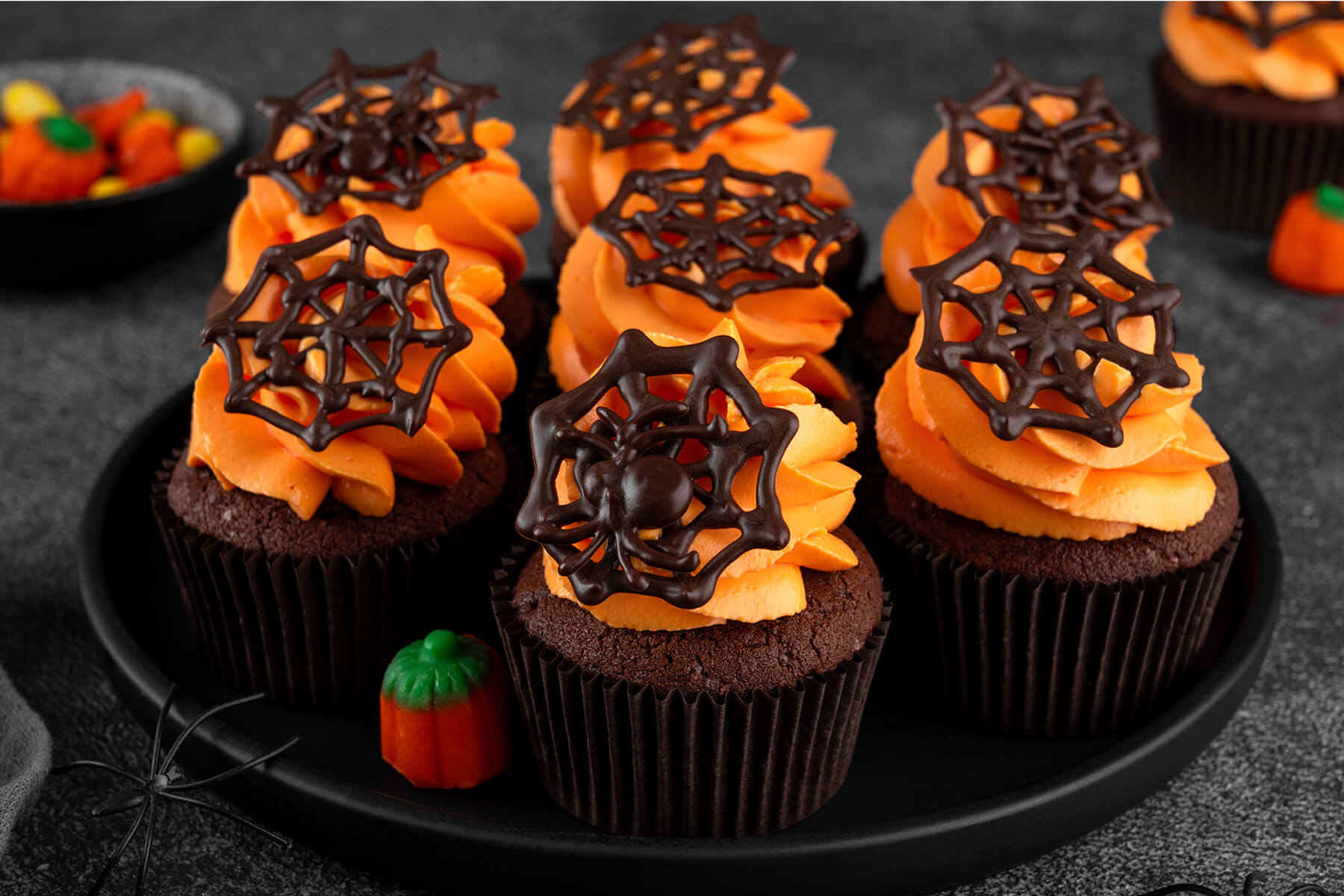 Mehrere Halloween-Schoko-Cupcakes mit oranger Buttercreme und dunklen Schoko-Spinnennetzen auf einem runden schwarzen Teller, der vordere mit einer Schoko-Spinne, dazu eine Kürbis-Zuckerfigur und eine schwarze Deko-Spinne am Tellerrand