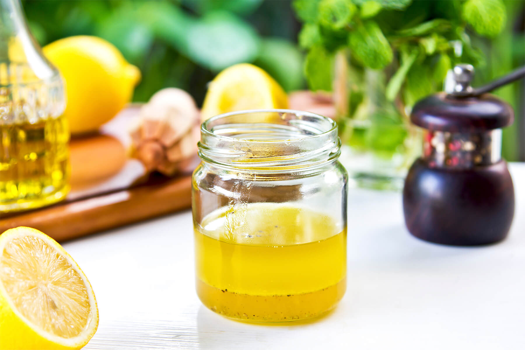 Kleines Glas Honig-Zitronen-Vinaigrette vor einem Hintergrund aus frischer Minze, mit Zitronen und einer Pfeffermühle