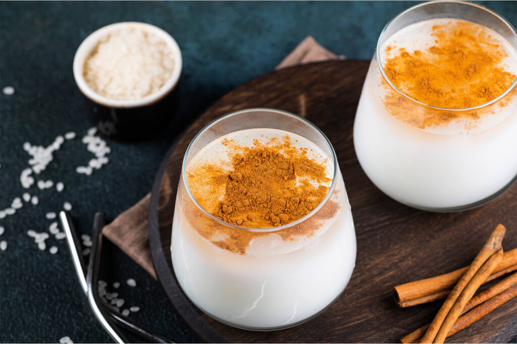 Zwei Gläser Horchata de Arroz mit gemahlenem Zimt obenauf, daneben Reis und Zimtstangen