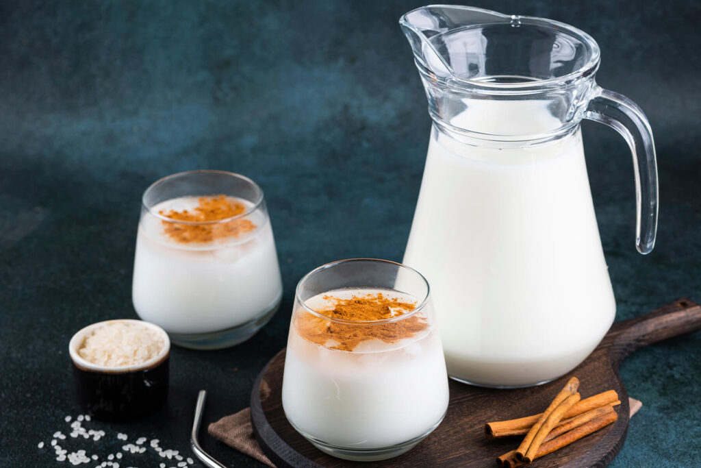 Glaskanne und zwei Gläser Horchata mit Zimt, Reiskörner und Zimtstangen auf dunklem Untergrund