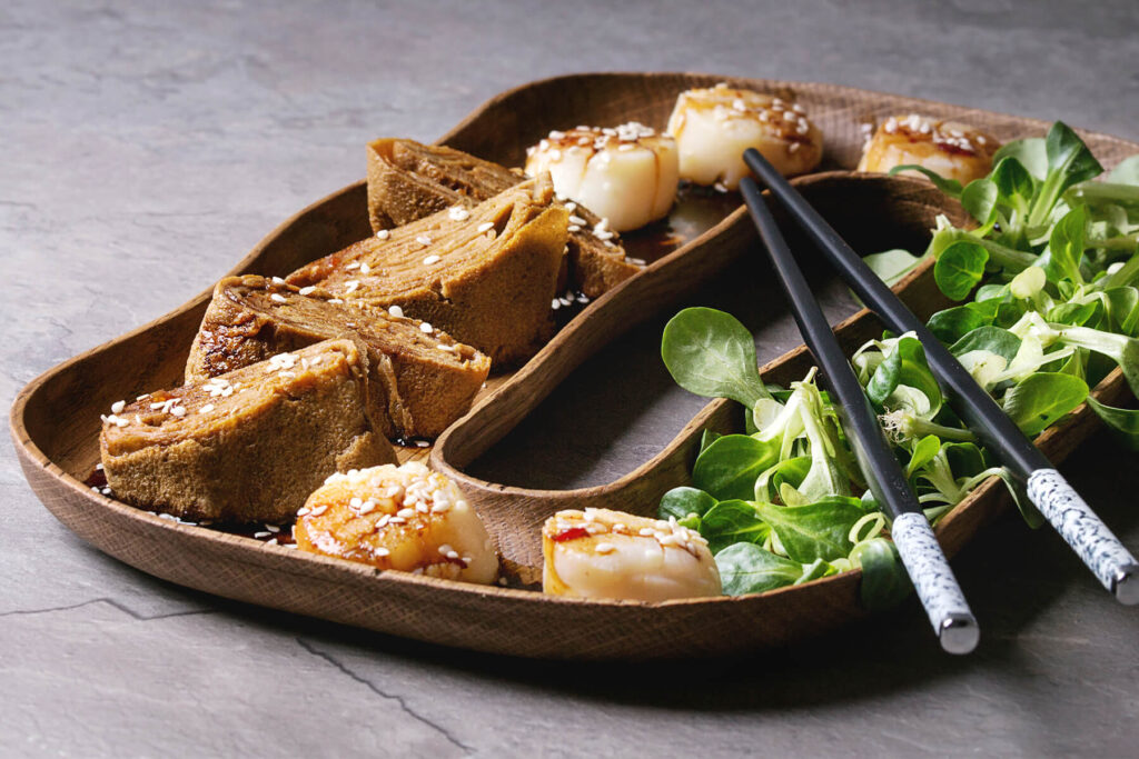 Zweigeteiltes ovales Holztablett aus der Nähe: fünf gebratene Jakobsmuscheln mit hell gebräunter Oberseite unter dunkler Sojaglasur und weißem Sesam, vier Scheiben nussbraunes Tamagoyaki-Rollomelett mit sichtbaren Spiralschichten, daneben Feldsalat und Stäbchen auf grauem Stein