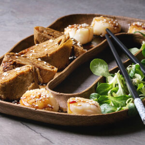 Zweigeteiltes ovales Holztablett aus der Nähe: fünf gebratene Jakobsmuscheln mit hell gebräunter Oberseite unter dunkler Sojaglasur und weißem Sesam, vier Scheiben nussbraunes Tamagoyaki-Rollomelett mit sichtbaren Spiralschichten, daneben Feldsalat und Stäbchen auf grauem Stein
