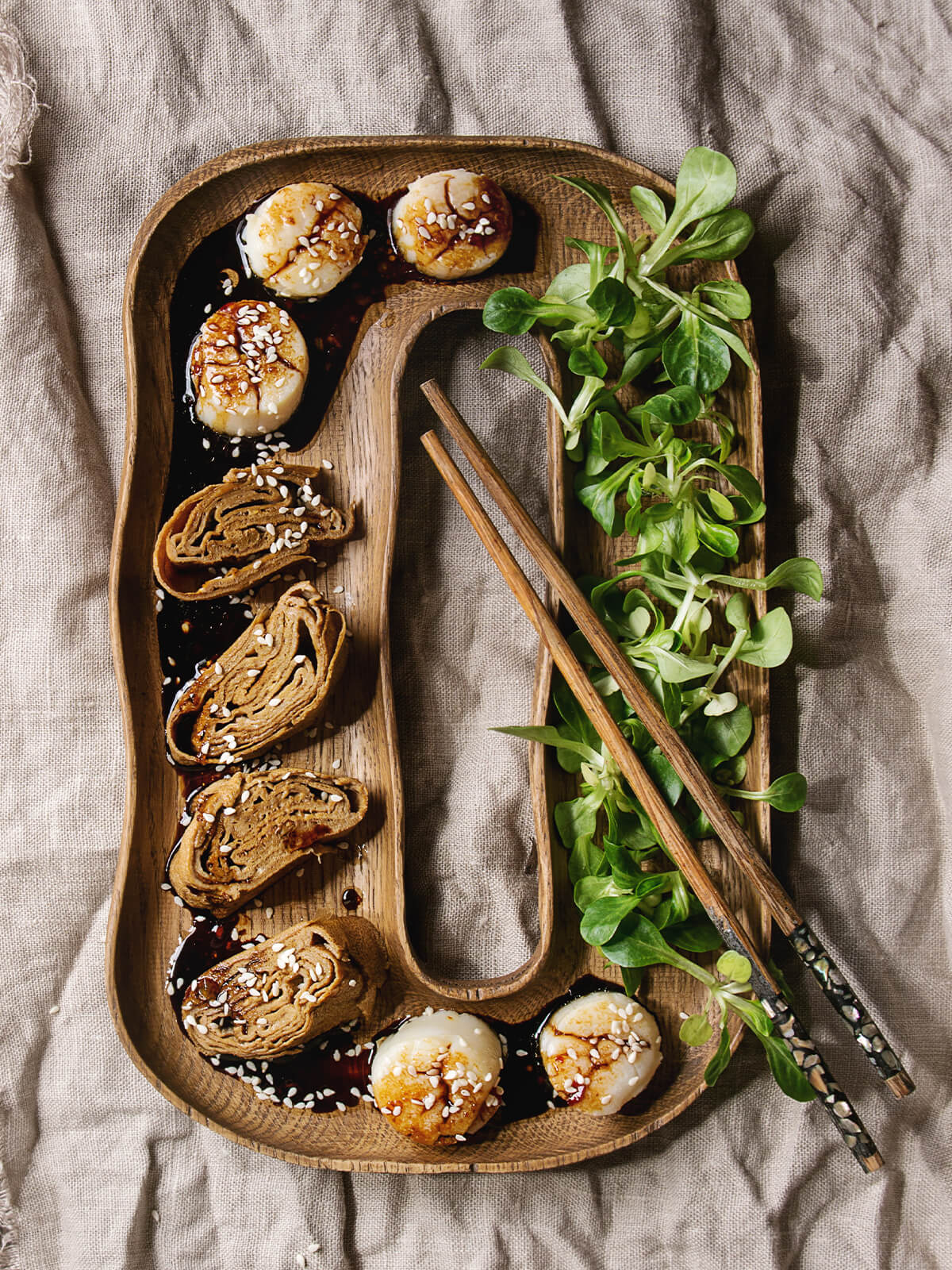 Japanische Vorspeisenplatte von oben auf hellem Leinentuch: geschwungenes Holztablett mit fünf gebratenen Jakobsmuscheln in dunkler Sojaglasur und weißem Sesam, vier Scheiben nussbraunem Tamagoyaki mit feinen Spiralschichten, Feldsalat in eigener Bahn und hellen Holzstäbchen