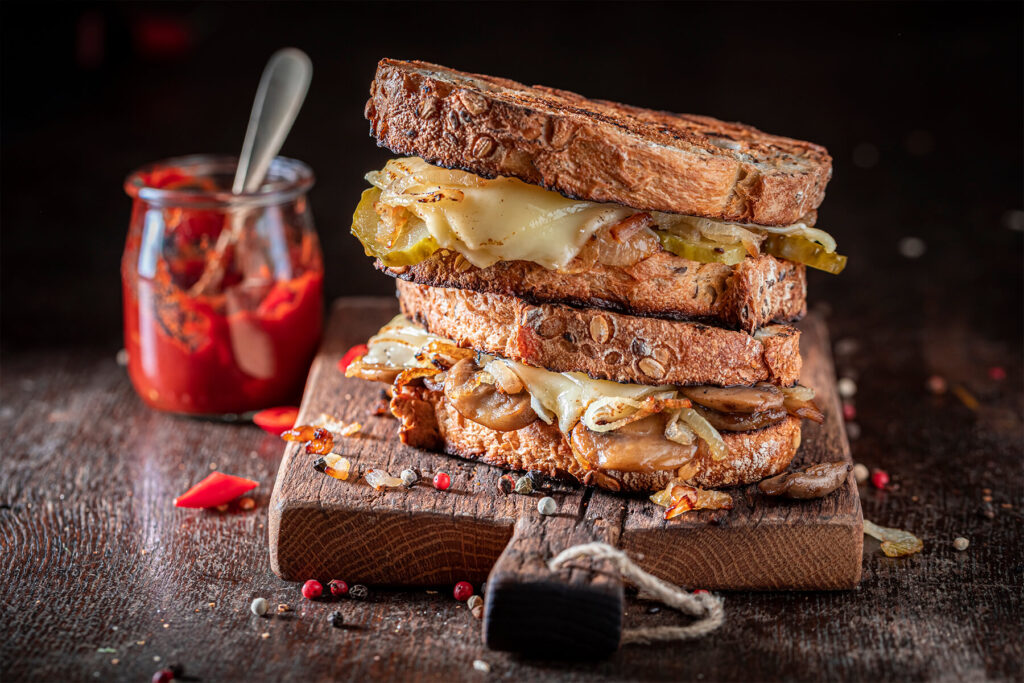 Geröstetes Käse-Sandwich mit geschmolzenem Käse, Champignons, Zwiebeln und Gewürzgurken, gestapelt auf einem Holzbrett
