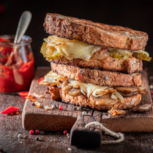 Geröstetes Käse-Sandwich mit geschmolzenem Käse, Champignons, Zwiebeln und Gewürzgurken, gestapelt auf einem Holzbrett