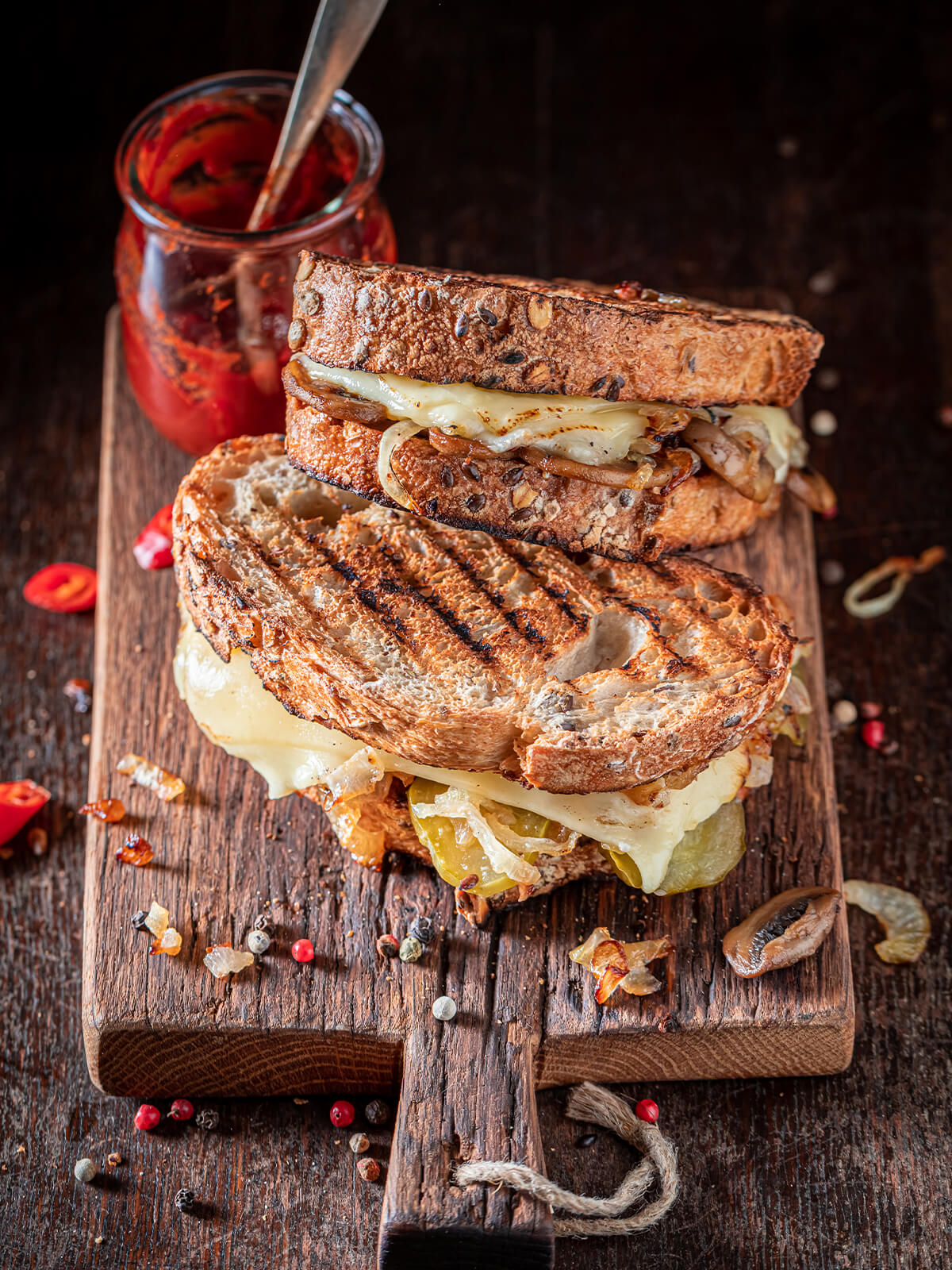 "Geröstetes Käse-Sandwich mit Pilzen und Gewürzgurken auf dunklem Untergrund neben einem Glas mit rotem Relish"