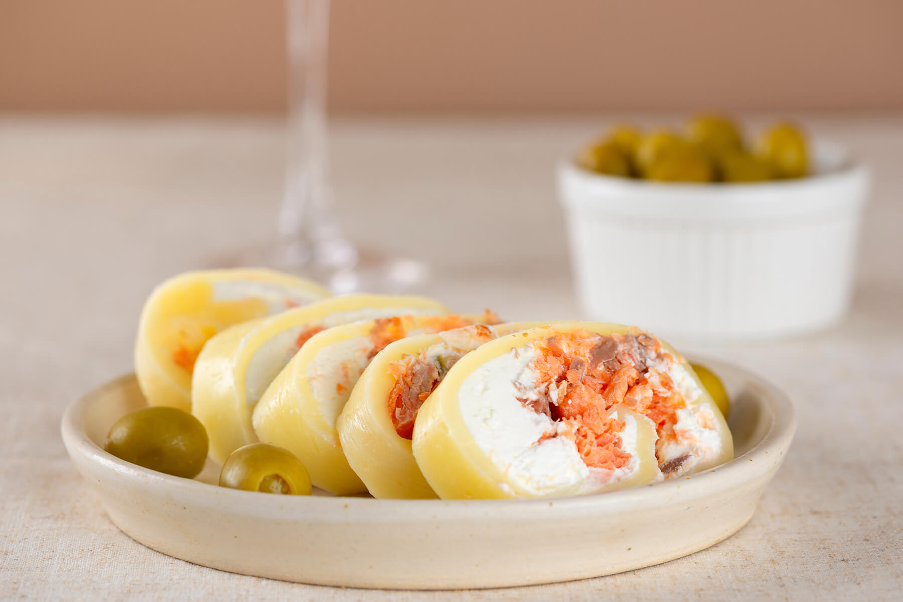 In Scheiben geschnittene Käserolle mit Lachs und Frischkäse auf einem hellen Teller, die dünne gelbe Käsehülle umschließt eine weiße Füllung mit zerpflücktem Lachs und Oliven, daneben grüne Oliven und ein Glas Weißwein