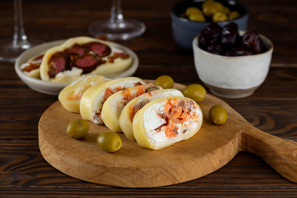 Herzhafte Käserolle mit Lachs und Frischkäse in Scheiben auf einem Holzbrett, garniert mit grünen Oliven, Datteln und gefriergetrockneten Erdbeeren, daneben ein Glas Weißwein