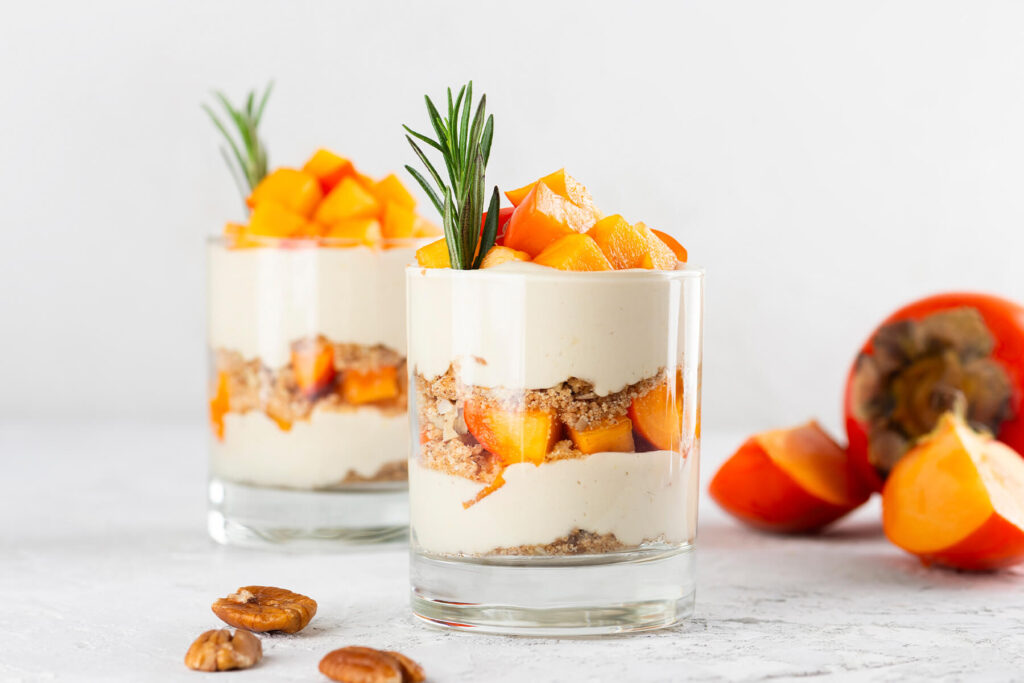 Zwei Gläser veganes Kaki-Parfait mit Schichten aus aufgeschlagener Kokoscreme, Pekan-Bröseln und leuchtend orangefarbenen Kaki-Würfeln, garniert mit Rosmarin, daneben ganze Kaki und geröstete Pekannüsse auf hellem Tisch