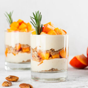 Zwei Gläser veganes Kaki-Parfait mit Schichten aus aufgeschlagener Kokoscreme, Pekan-Bröseln und leuchtend orangefarbenen Kaki-Würfeln, garniert mit Rosmarin, daneben ganze Kaki und geröstete Pekannüsse auf hellem Tisch