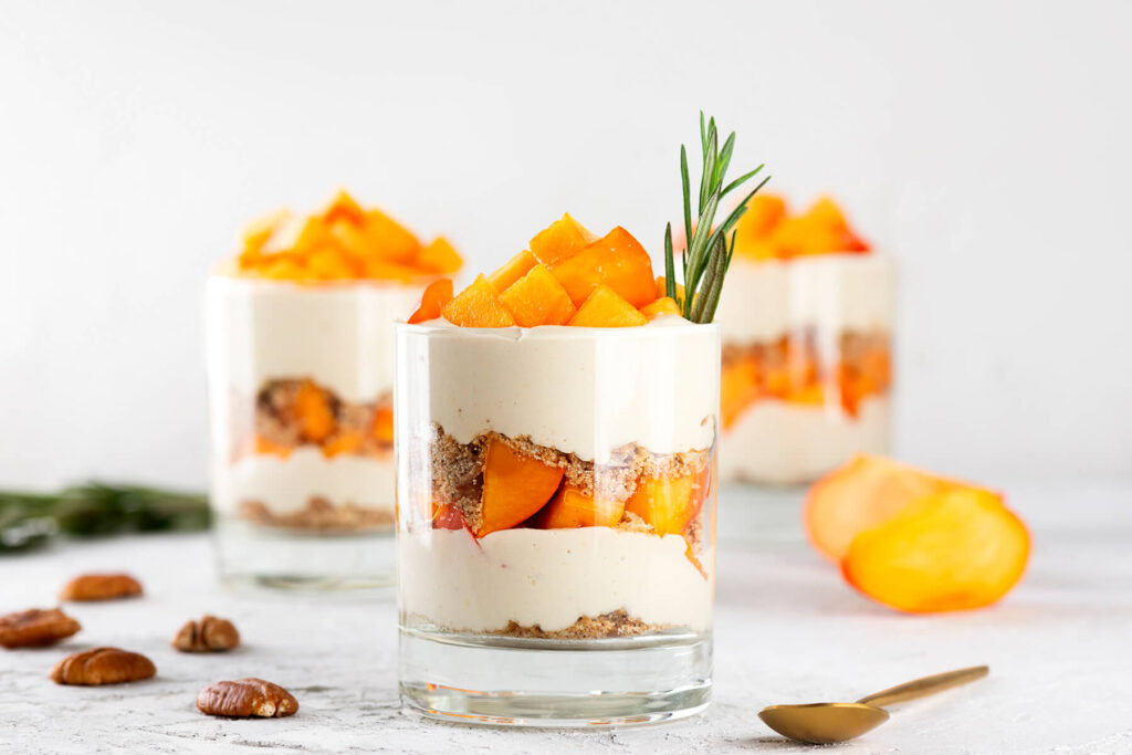 Ein Glas veganes Kaki-Parfait von vorn mit klar sichtbaren Schichten aus Kokoscreme, Pekan-Bröseln und Kaki, obenauf Kaki-Würfel und ein Rosmarinzweig, daneben ein goldener Löffel und geröstete Pekannüsse