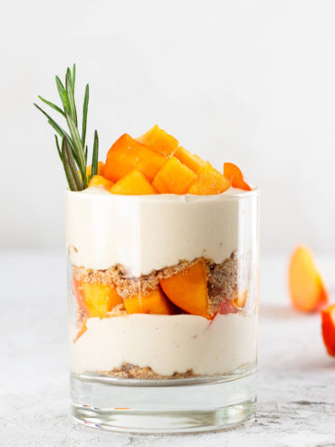 Ein Glas veganes Kaki-Parfait mit Schichten aus Pekan-Bröseln, luftig aufgeschlagener Kokoscreme und leuchtend orangefarbenen Kaki-Würfeln, garniert mit einem Rosmarinzweig, vor hellem Hintergrund