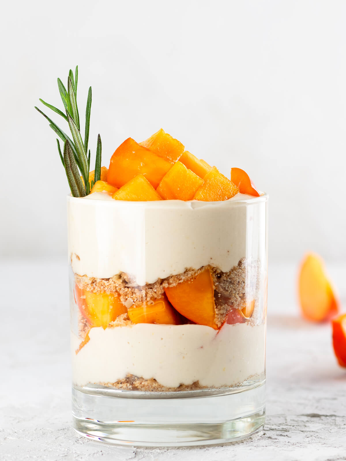 Ein Glas veganes Kaki-Parfait mit Schichten aus Pekan-Bröseln, luftig aufgeschlagener Kokoscreme und leuchtend orangefarbenen Kaki-Würfeln, garniert mit einem Rosmarinzweig, vor hellem Hintergrund
