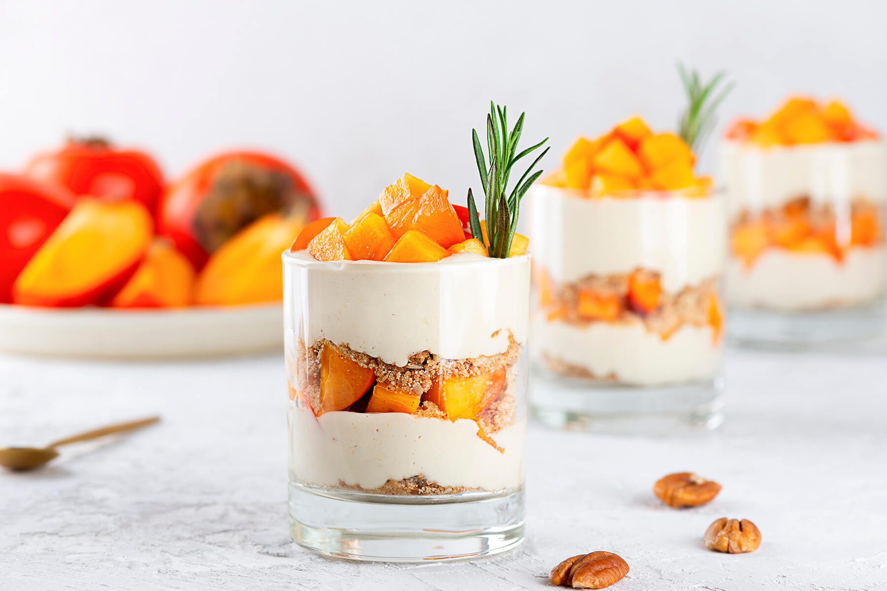 Drei Gläser veganes Kaki-Parfait nebeneinander mit deutlich sichtbaren Schichten aus Kokoscreme, Pekan-Bröseln und Kaki, jeweils mit Rosmarin garniert, im Hintergrund ein Teller mit frischen Kaki und geröstete Pekannüsse ringsum