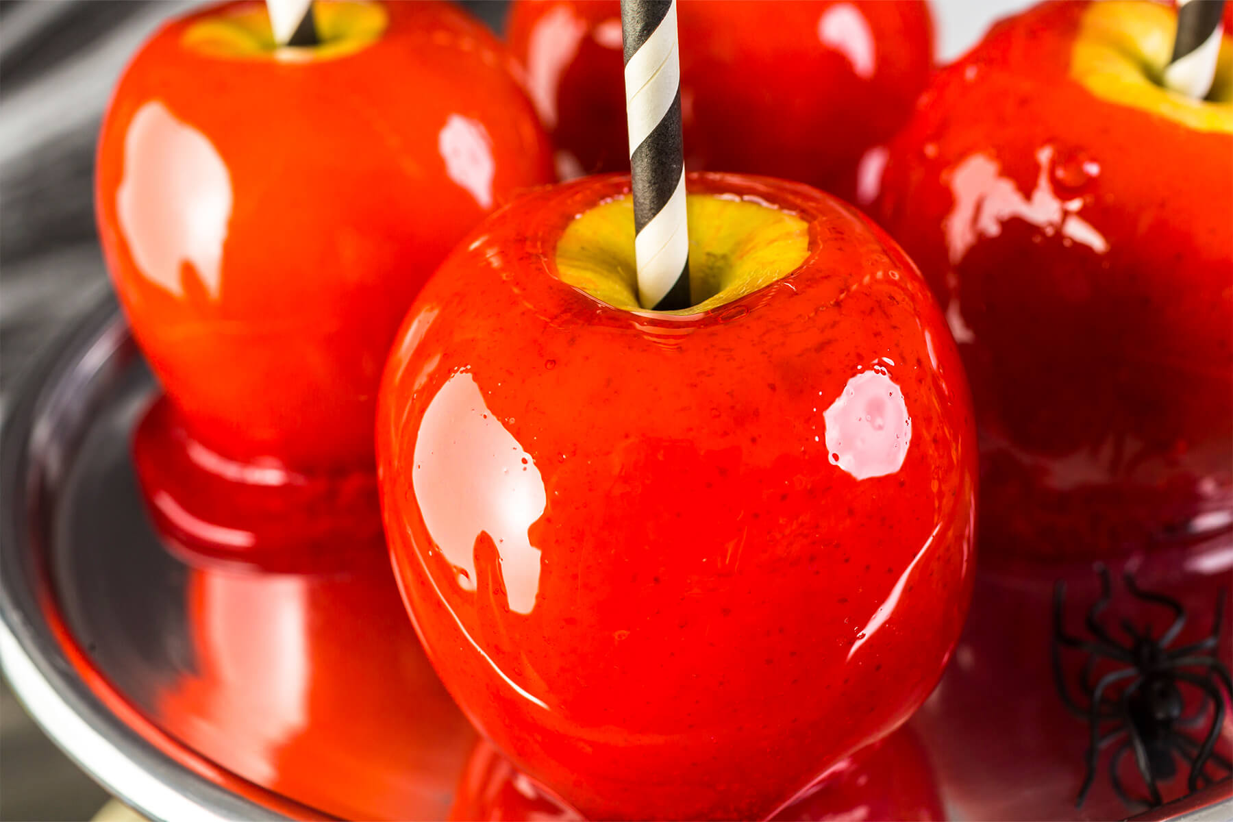 Nahaufnahme mehrerer roter Liebesäpfel auf einer Metallplatte, die durchscheinende Zuckerglasur spiegelt das Licht in klar abgegrenzten hellen Flecken und läuft am Fuß zu einem dünnen glasigen Kragen aus; oben um die schwarz-weiß gestreiften Papierstrohhalme liegt in der Stielmulde ein Ring blanker gelber Apfelschale frei, rechts unten eine schwarze Deko-Spinne aus Kunststoff