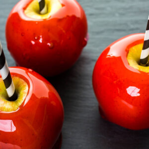 Drei rote Liebesäpfel von schräg oben auf dunkler Schieferplatte, die durchscheinende Zuckerglasur glatt und spiegelnd mit einzelnen Luftbläschen darin; der Überzug reicht bis an den Rand der Stielmulde, in der um den schwarz-weiß gestreiften Papierstrohhalm ein Ring blanker gelber Apfelschale frei liegt, an den Füßen sind erstarrte Glasurränder zu sehen