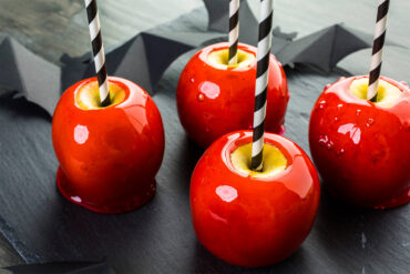 Vier rote Liebesäpfel an schwarz-weiß gestreiften Papierstrohhalmen auf einer dunklen Schieferplatte, dazwischen ausgeschnittene Fledermäuse aus schwarzem Papier; die Zuckerglasur ist hochglänzend und durchscheinend rot, oben um jeden Halm liegt ein Ring gelber Apfelschale frei, an den Füßen ist die Glasur zu flachen Kragen ausgelaufen