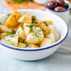 Weiße Emailschale mit dunkelblauem Rand, schräg von der Seite fotografiert, gefüllt mit geviertelten kleinen Kartoffeln mit teilweise hellbrauner Schale, ölglänzend, bestreut mit feinen grünen Dillspitzen und einzelnen ganzen grünen Kapernknospen; im unscharfen Hintergrund ein kleines Emailschälchen mit dunklen Oliven, ein Holzbrett und ein hellblaues Leinentuch
