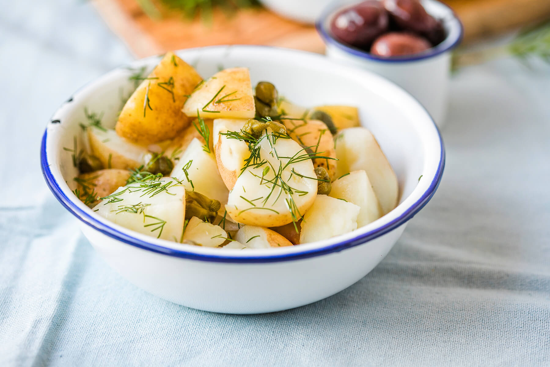 Weiße Emailschale mit dunkelblauem Rand, schräg von der Seite fotografiert, gefüllt mit geviertelten kleinen Kartoffeln mit teilweise hellbrauner Schale, ölglänzend, bestreut mit feinen grünen Dillspitzen und einzelnen ganzen grünen Kapernknospen; im unscharfen Hintergrund ein kleines Emailschälchen mit dunklen Oliven, ein Holzbrett und ein hellblaues Leinentuch