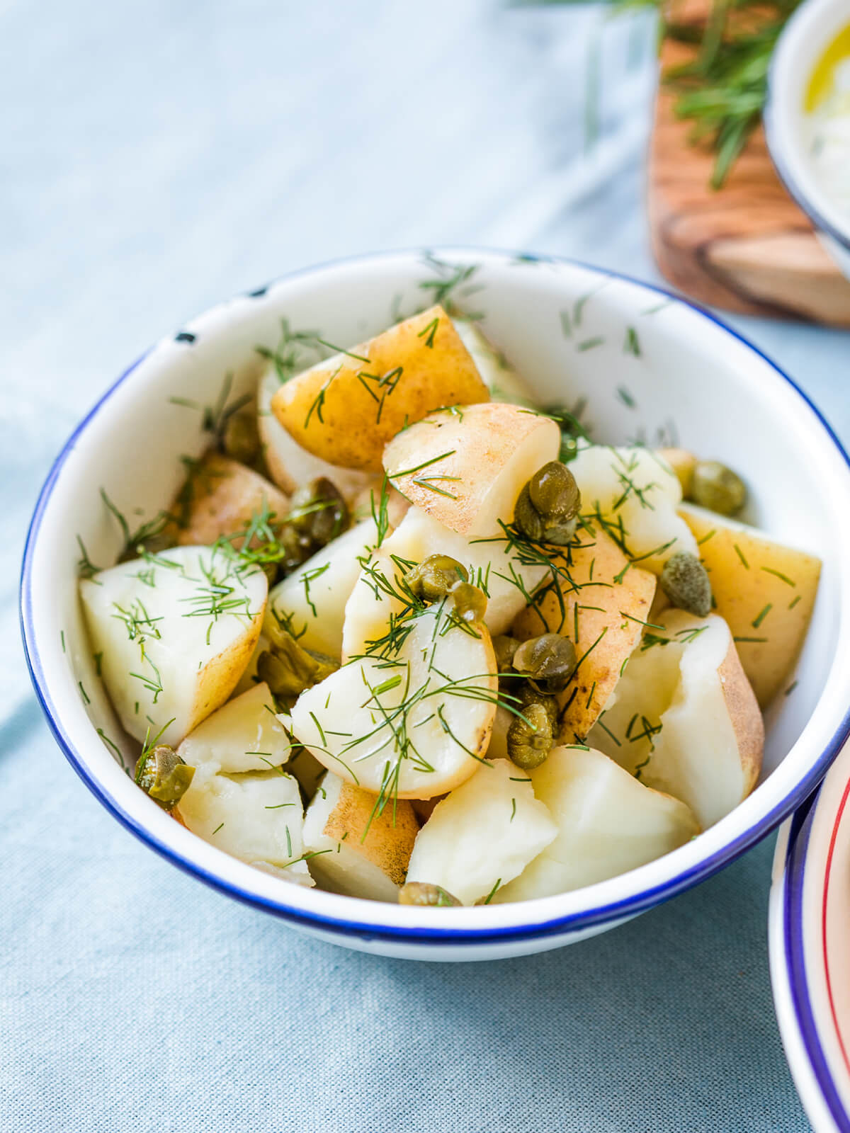 Nahaufnahme schräg von oben in eine weiße Emailschale mit dunkelblauem Rand: geviertelte kleine Kartoffeln mit teilweise hellbrauner Schale, ölglänzend, dicht bestreut mit feinen Dillspitzen, dazwischen ganze grüne Kapernknospen; im Hintergrund unscharf ein Holzbrett mit Rosmarinzweigen und eine helle Schale, darunter ein hellblaues Leinentuch