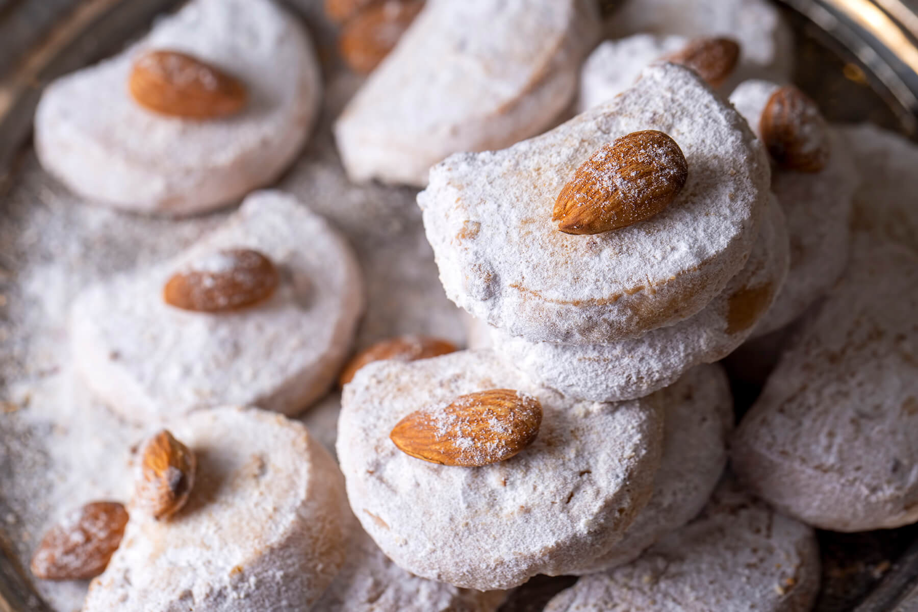 Mehrere Kavala Cookies in Halbmondform auf einer silbernen Platte, jeder mit einer ganzen Mandel und dicker Puderzuckerschicht