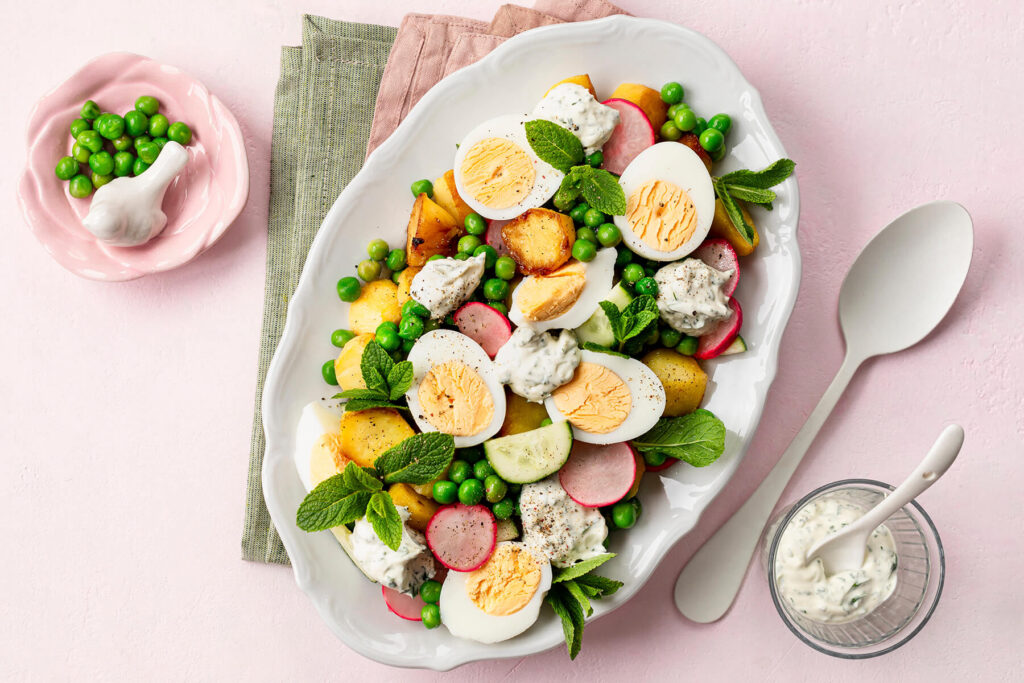 Knuspriger Kartoffel-Ei-Salat auf einer weißen Platte von oben, mit goldgelben, an den Kanten gebratenen Kartoffeln, halbierten Eiern, grünen Erbsen, Radieschen, Gurke, Minze und Klecksen cremigem Joghurt-Kräuter-Dressing, daneben ein Glas Dressing und eine Schale Erbsen
