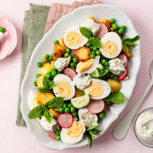 Knuspriger Kartoffel-Ei-Salat auf einer weißen Platte von oben, mit goldgelben, an den Kanten gebratenen Kartoffeln, halbierten Eiern, grünen Erbsen, Radieschen, Gurke, Minze und Klecksen cremigem Joghurt-Kräuter-Dressing, daneben ein Glas Dressing und eine Schale Erbsen